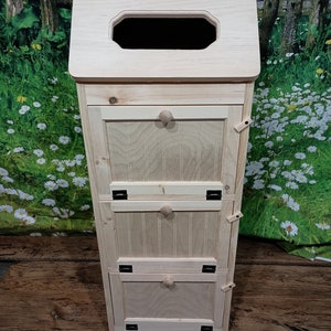 May include: A wooden three-tiered storage cabinet with a hinged top and three doors. Each door has a small knob and is accented with black hinges. The cabinet is made of light wood and has a rustic, farmhouse style.