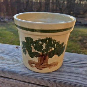 May include: A cream-colored ceramic planter with a green band around the top and a hand-painted design of a brown tree with green leaves.