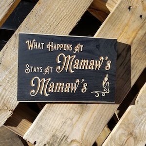 May include: A black wooden sign with gold lettering that reads "What happens at Mamaw's stays at Mamaw's".