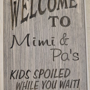 May include: A wooden sign with the text "Welcome to Mimi & Pa's Kids Spoiled While You Wait!" in black lettering on a gray background.