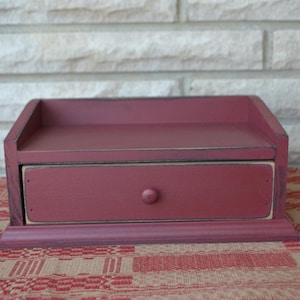 May include: A small, red wooden box with a single drawer. The box has a distressed paint finish and a simple design.