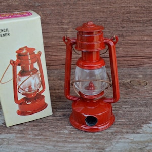 May include: A red metal pencil sharpener shaped like a vintage kerosene lantern. The sharpener has a clear plastic window and a metal handle. The box reads "Pencil Sharpener".