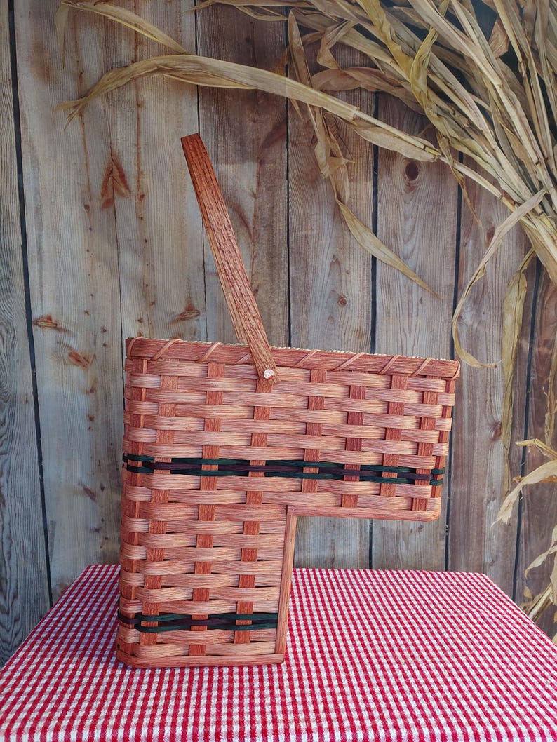 May include: A brown wicker basket with a wooden handle and black and green trim. The basket is sitting on a red and white checkered tablecloth.