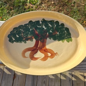 May include: A yellow oval ceramic serving tray with a green tree painted on it. The tree has brown roots and a small bird perched on a branch.