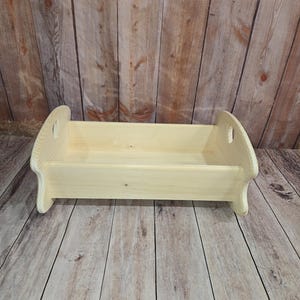 May include: A natural wood shelf with a rectangular base and curved sides. The shelf has two handles and is made of unfinished wood. The background is a wooden surface.