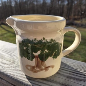 May include: A cream-colored ceramic pitcher with a green tree design. The pitcher has a brown handle and a wide mouth.