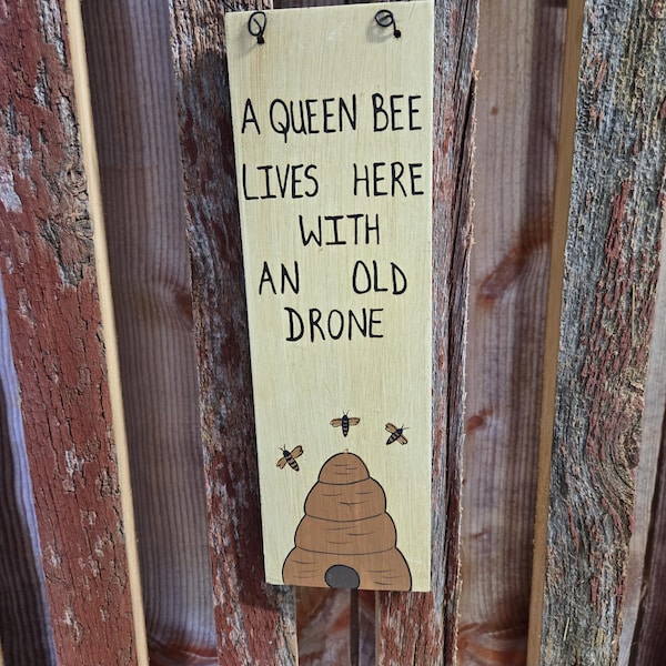 Painted wood sign "A Queen Bee Lives Here With Her Old Drone". It has a bee hive and 3 bees