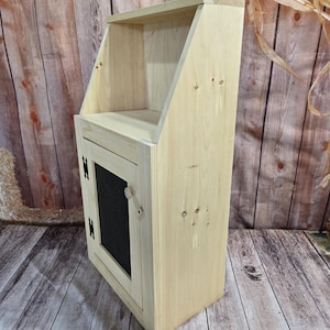 May include: A small, unfinished wooden cabinet with a single door and a shelf. The door has a black painted panel and a simple knob.