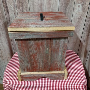 May include: A rustic wooden box with a red and white painted finish. The box has a hinged lid with a black metal handle. The box is sitting on a red and white checkered tablecloth.