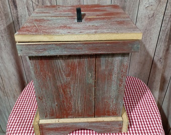 May include: A rustic wooden box with a red and white painted finish. The box has a hinged lid with a black metal handle. The box is sitting on a red and white checkered tablecloth.
