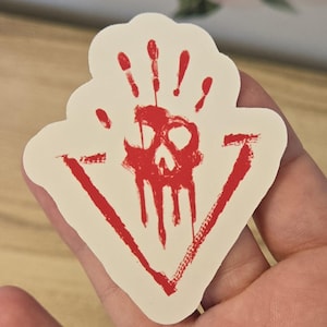 May include: A sticker with a red skull and handprint design. The skull is in the center, with a handprint above it and dripping blood. The design is enclosed in a red triangle. The sticker is on a cream-colored background.