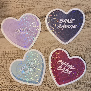 May include: Four heart-shaped stickers with holographic glitter accents. Each sticker has a different color and text: purple with "SHAR SYMPATHIZER", dark blue with "BANE BADDIE", light blue with "MYRRUL MANIAC", and red with "BHAAL BABE".