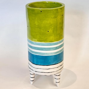 May include: A tall, ceramic vase with a vibrant design. The top section is lime green with black dots, transitioning to white and blue stripes, and a solid blue band. The vase rests on three white legs with black stripes.