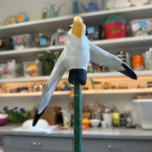 Op de afbeelding: Handgemaakte vogelsculptuur met uitgestrekte vleugels, met een wit lichaam, gele snavel en zwarte vleugeltips. De vogel is gemonteerd op een groene paal met een zwarte basis, tegen een vage achtergrond van planken.