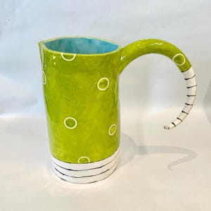 May include: A lime green ceramic pitcher featuring white polka dots on the body, a light blue interior, and a curved handle with black and white stripes. The base of the pitcher is decorated with horizontal black and white stripes.