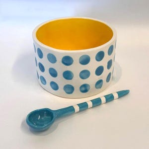 May include: A ceramic bowl with a yellow interior and white exterior decorated with blue polka dots. A matching blue spoon with white stripes rests beside the bowl. The spoon has a small, rounded bowl.