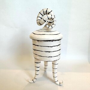 May include: A white ceramic lidded jar with black horizontal stripes. The lid features a prominent, spiraled handle. The jar is supported by three slender, curved legs that taper to points, all decorated with matching black horizontal stripes.