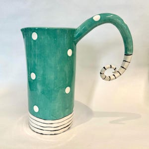 May include: A tall ceramic pitcher with a glossy teal glaze and white polka dots. The base features horizontal black stripes, and the handle is a long, curved teal extension that ends in a spiral with black and white striped detailing.
