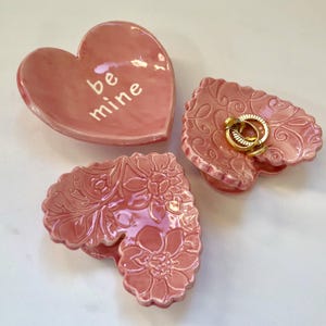 May include: Three pink heart-shaped ceramic dishes. One dish has the words "be mine" written on it. The other two dishes have a floral design.