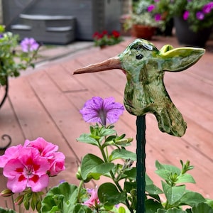 May include: A ceramic hummingbird garden stake with a green and brown glaze. The bird has a long, brown beak and is mounted on a green metal stake. Pink and purple flowers are in the foreground.