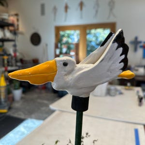 May include: A ceramic pelican garden stake with a white body, yellow beak, and black wing accents. The pelican is mounted on a green metal rod with a black base. The bird is in a flying pose.