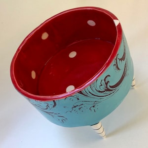 Puede incluir: Un cuenco de cerámica rojo con lunares blancos y un diseño floral azul y blanco. El cuenco tiene tres patas blancas.