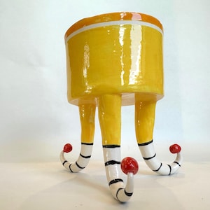 May include: A yellow ceramic planter with a glossy finish and a white rim. The planter is supported by three legs that transition from yellow to white with black horizontal stripes, ending in curled tips topped with small, round red spheres.