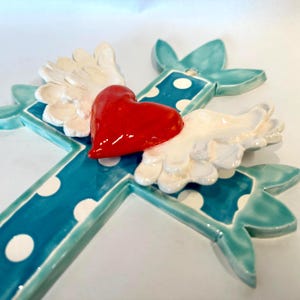 May include: A ceramic cross wall hanging featuring a glossy teal blue finish with white polka dots. The center of the cross displays a three-dimensional bright red heart with sculpted white wings extending outward on both sides.