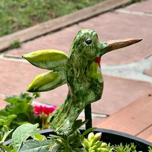 May include: A green ceramic hummingbird garden stake with yellow-tipped wings and a brown beak. The bird has a red patch on its chest and is mounted on a dark green metal stake. The hummingbird is placed in a flower pot with green foliage and pink flowers.
