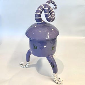 May include: A lavender ceramic lidded jar with three legs and a tall, spiraling handle. The jar features white polka dots and small green floral accents. The legs and the handle are decorated with horizontal white stripes on a lavender base.