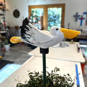 May include: A white ceramic pelican garden stake with a black and white wing and a yellow beak. The pelican is mounted on a green metal stake, perfect for adding a touch of coastal charm to any garden or outdoor space.