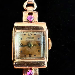 Könnte beinhalten: Eine Vintage-Zodiac-Armbanduhr mit goldfarbenem Gehäuse und Armband. Das Zifferblatt der Uhr ist hellbraun mit schwarzen Ziffern und Zeigern. Zwei kleine rosa Edelsteine ​​sind auf dem Armband eingesetzt.