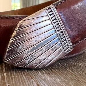 May include: A brown leather belt with a silver buckle. The buckle has a decorative design with a textured surface.