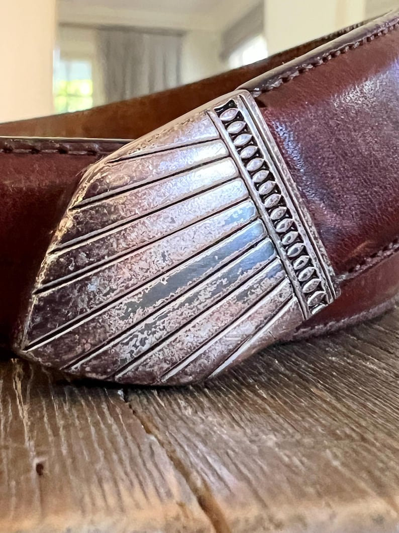 May include: A brown leather belt with a silver buckle. The buckle has a decorative design with a textured surface.