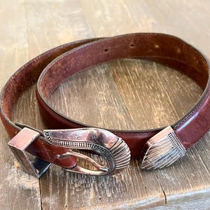 May include: A brown leather belt with a silver buckle featuring an intricate design. The buckle is a traditional western style.