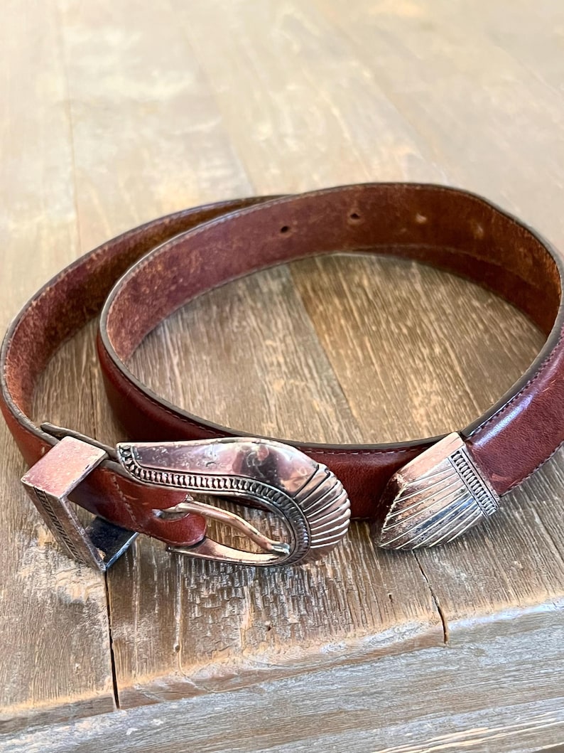 May include: A brown leather belt with a silver buckle featuring an intricate design. The buckle is a traditional western style.