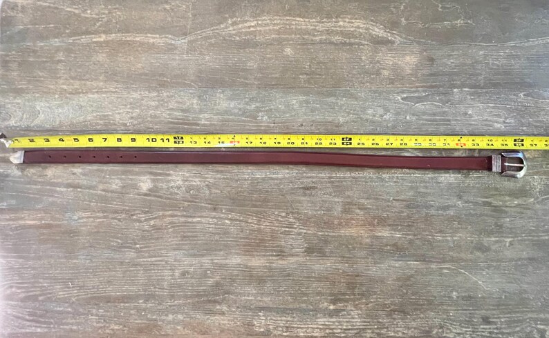 May include: A brown leather belt with a silver buckle. The belt is measured with a yellow tape measure, showing it is 39 inches long.