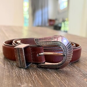 May include: A brown leather belt with a silver buckle. The buckle has a decorative design with a southwestern style.