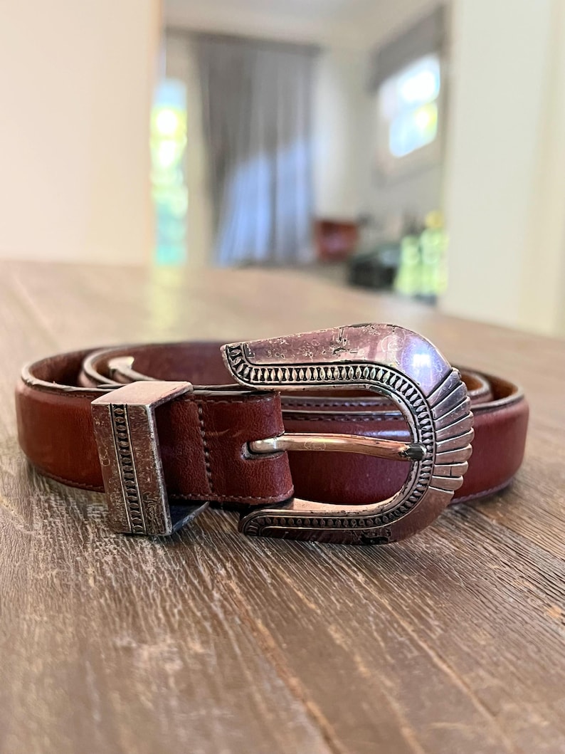 May include: A brown leather belt with a silver buckle. The buckle has a decorative design with a southwestern style.