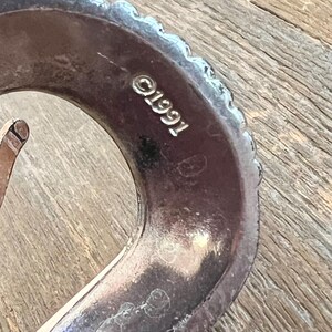 May include: Close-up of a silver belt buckle with a textured edge and the inscription "C1991".