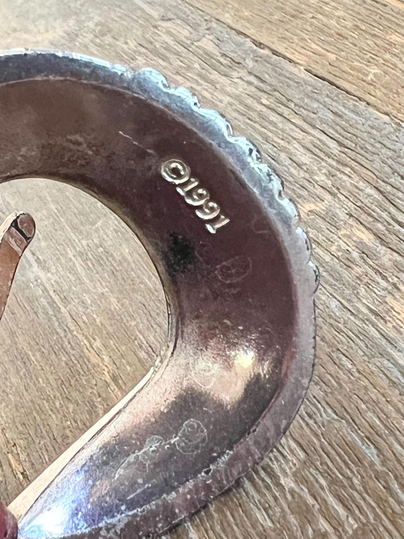 May include: Close-up of a silver belt buckle with a textured edge and the inscription "C1991".