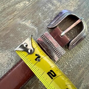 May include: A brown leather belt with a silver buckle. The buckle has a decorative design and is measured with a yellow tape measure.