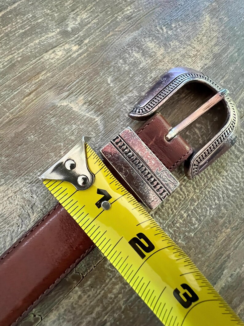 May include: A brown leather belt with a silver buckle. The buckle has a decorative design and is measured with a yellow tape measure.
