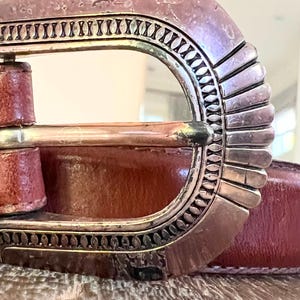 May include: Close-up of a brown leather belt with a silver buckle. The buckle has a decorative design with a scalloped edge.
