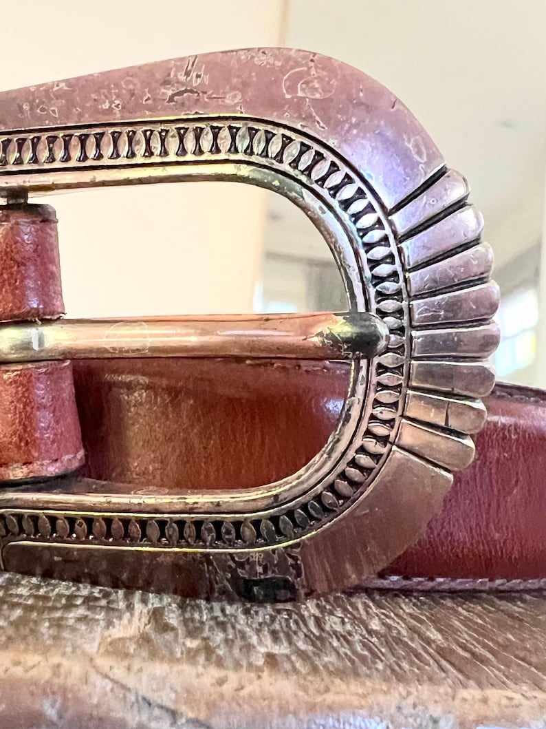 May include: Close-up of a brown leather belt with a silver buckle. The buckle has a decorative design with a scalloped edge.