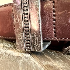May include: A brown leather belt with a silver buckle. The buckle has a decorative design with a repeating pattern of ovals.