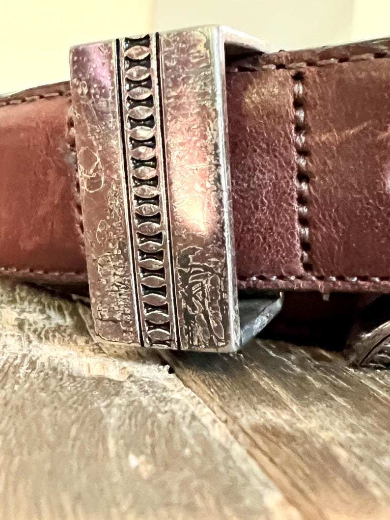May include: A brown leather belt with a silver buckle. The buckle has a decorative design with a repeating pattern of ovals.