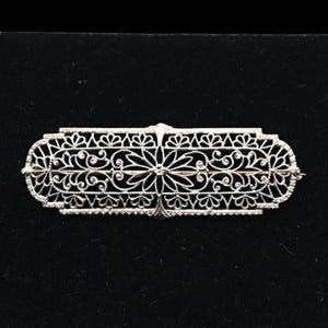 May include: A rectangular silver-toned metal brooch featuring an intricate openwork filigree design. The piece displays a symmetrical pattern of delicate scrolling vines, floral motifs, and geometric borders, creating a detailed lace-like appearance.