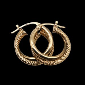 May include: A pair of gold hoop earrings featuring a twisted rope texture along the outer surface. The earrings are circular in shape with a hinged post closure mechanism. The metal surface is polished and reflective, showing a 10K stamp on the post.