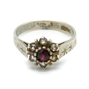 May include: A silver ring with a red gemstone surrounded by smaller white gemstones. The band is engraved with a pattern of stars.